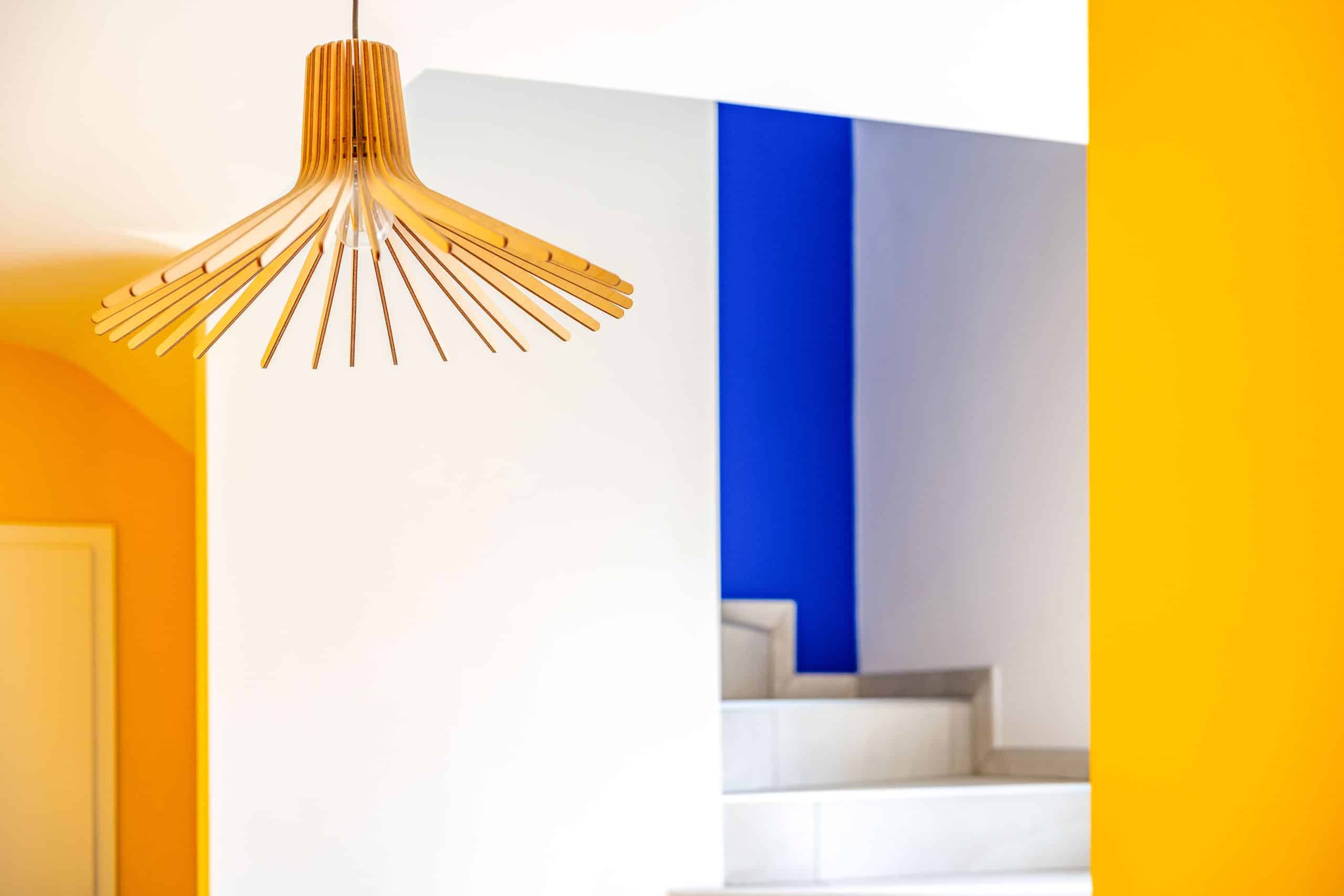 Suspension design en bois et jeu de couleurs vives dans une cage d’escalier rénovée — jaune, blanc et bleu électrique — lors de la rénovation d’une maison des années 2000 par Azai – Aléna Zuliani Azai architecture